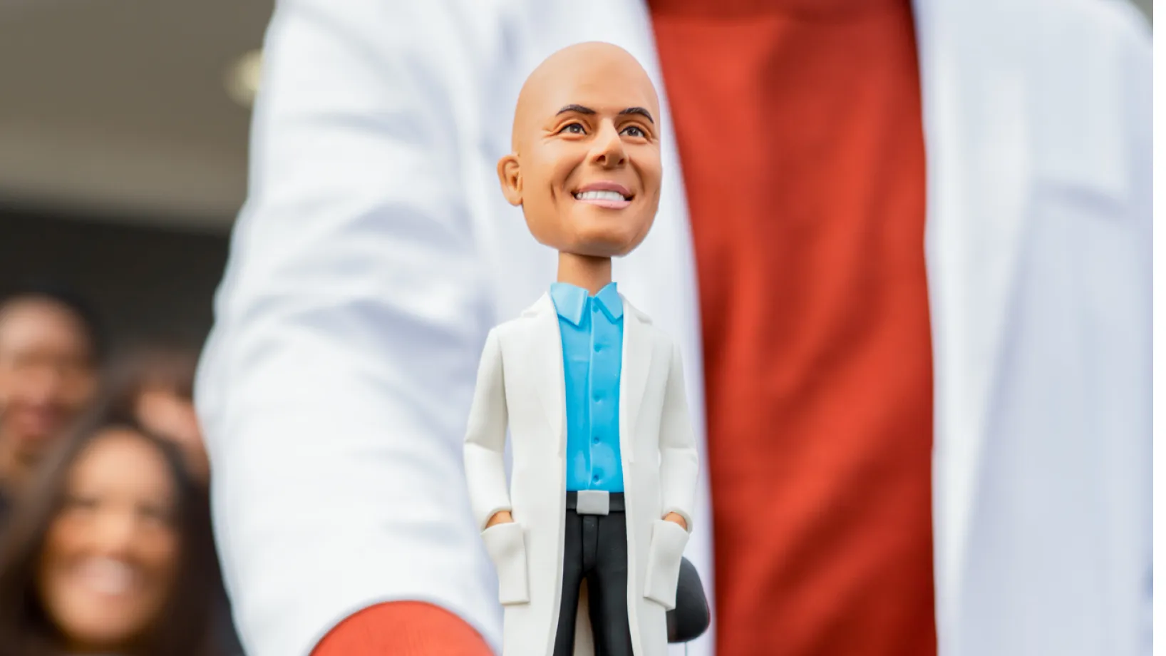 Person holding a smiling bobblehead figure dressed in a white coat and blue shirt.