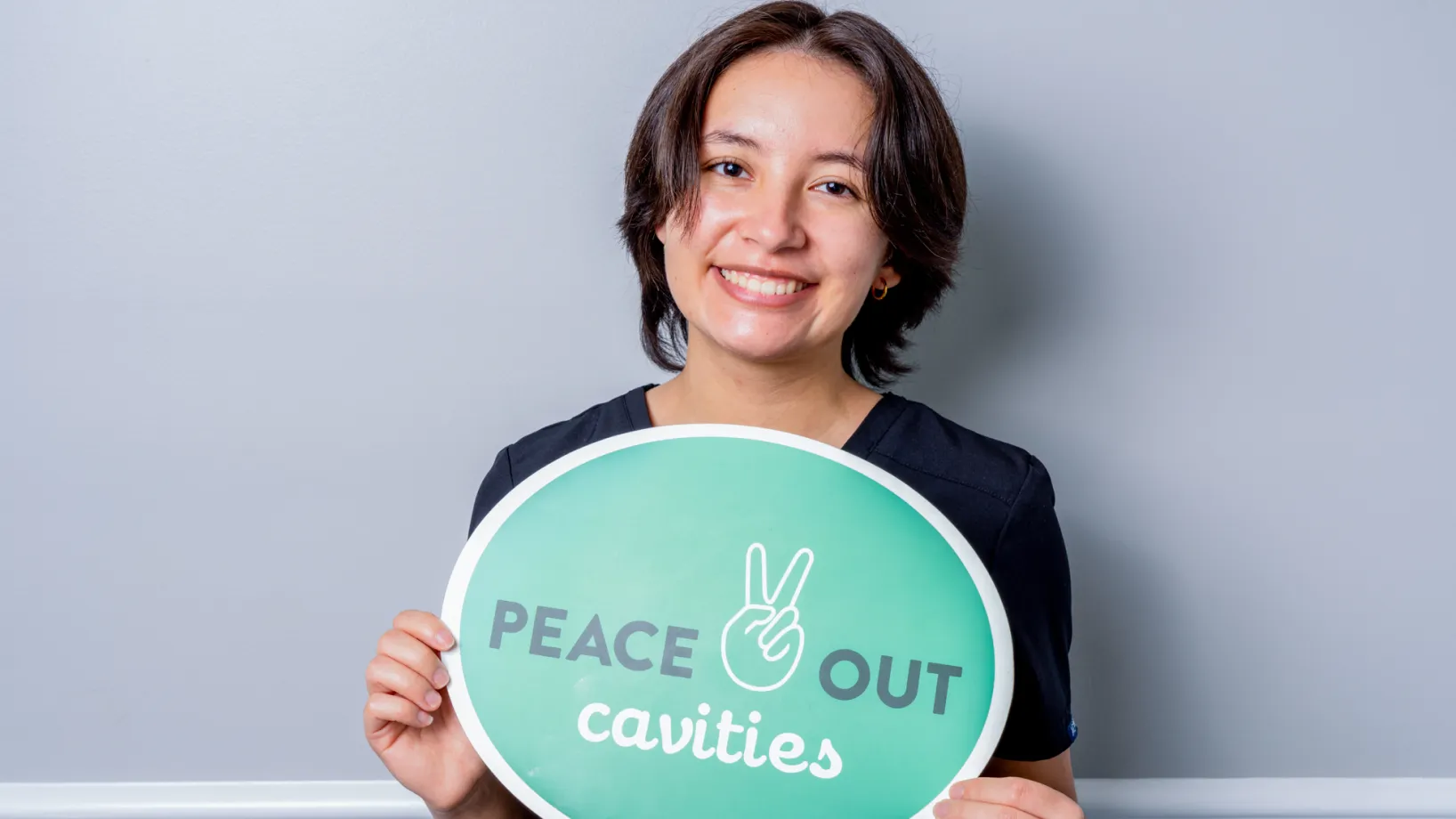 Smiling young person in dark clothing holding a green sign that says 'Peace Out cavities' with a peace hand symbol.