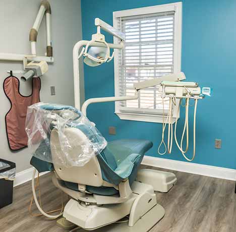 Modern dental examination chair with overhead light and dental tools in a clean room with blue accent wall and window blinds.