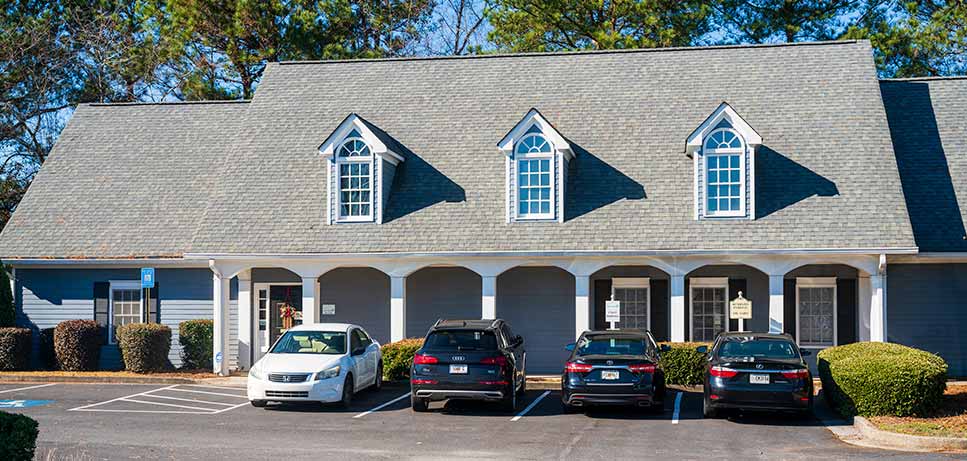 Front view of a light gray building with three dormer windows and four parked cars in front.