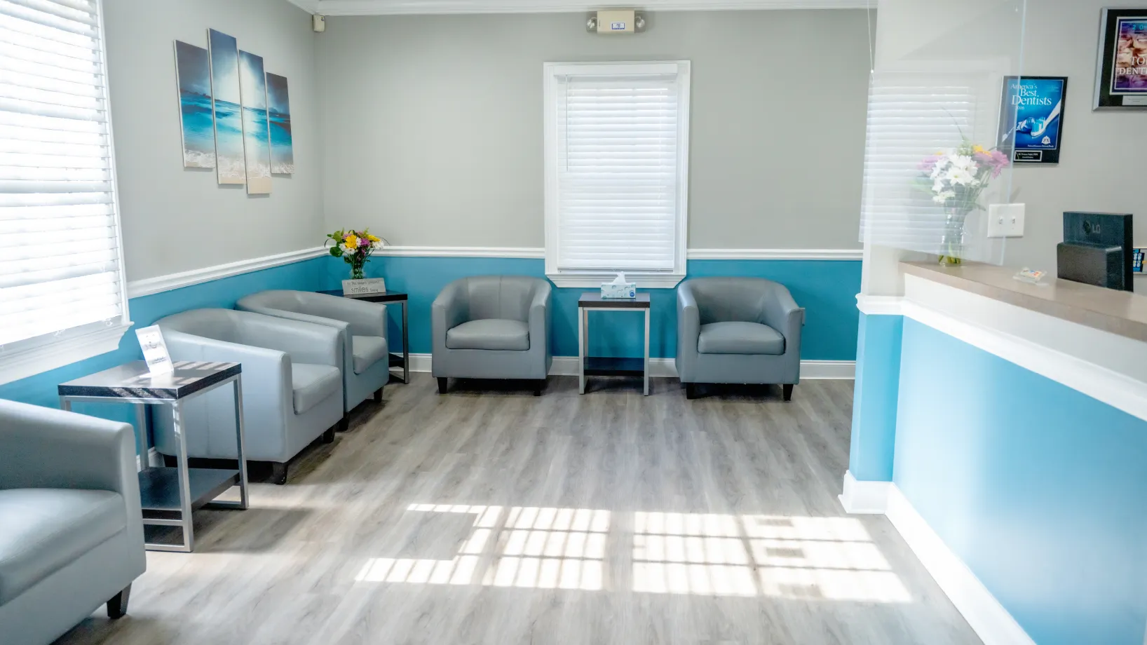 Modern waiting room with light gray armchairs, blue and gray walls, wood flooring, and a reception desk with flowers.