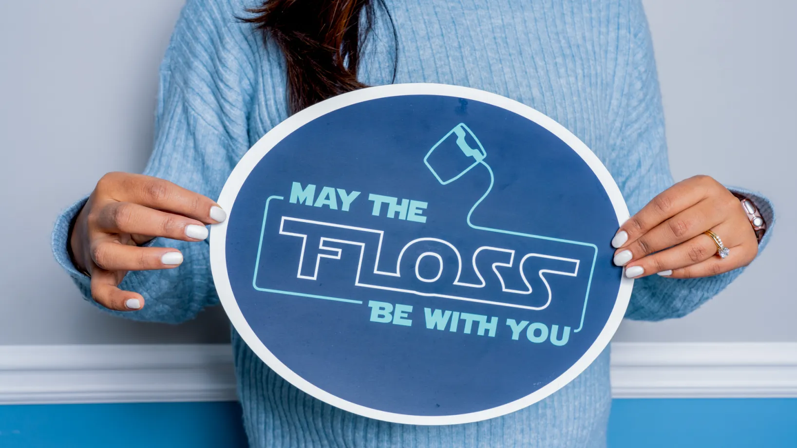 Person wearing a blue sweater holding a blue and white sign that reads 'May the Floss be with you' with a dental floss icon.