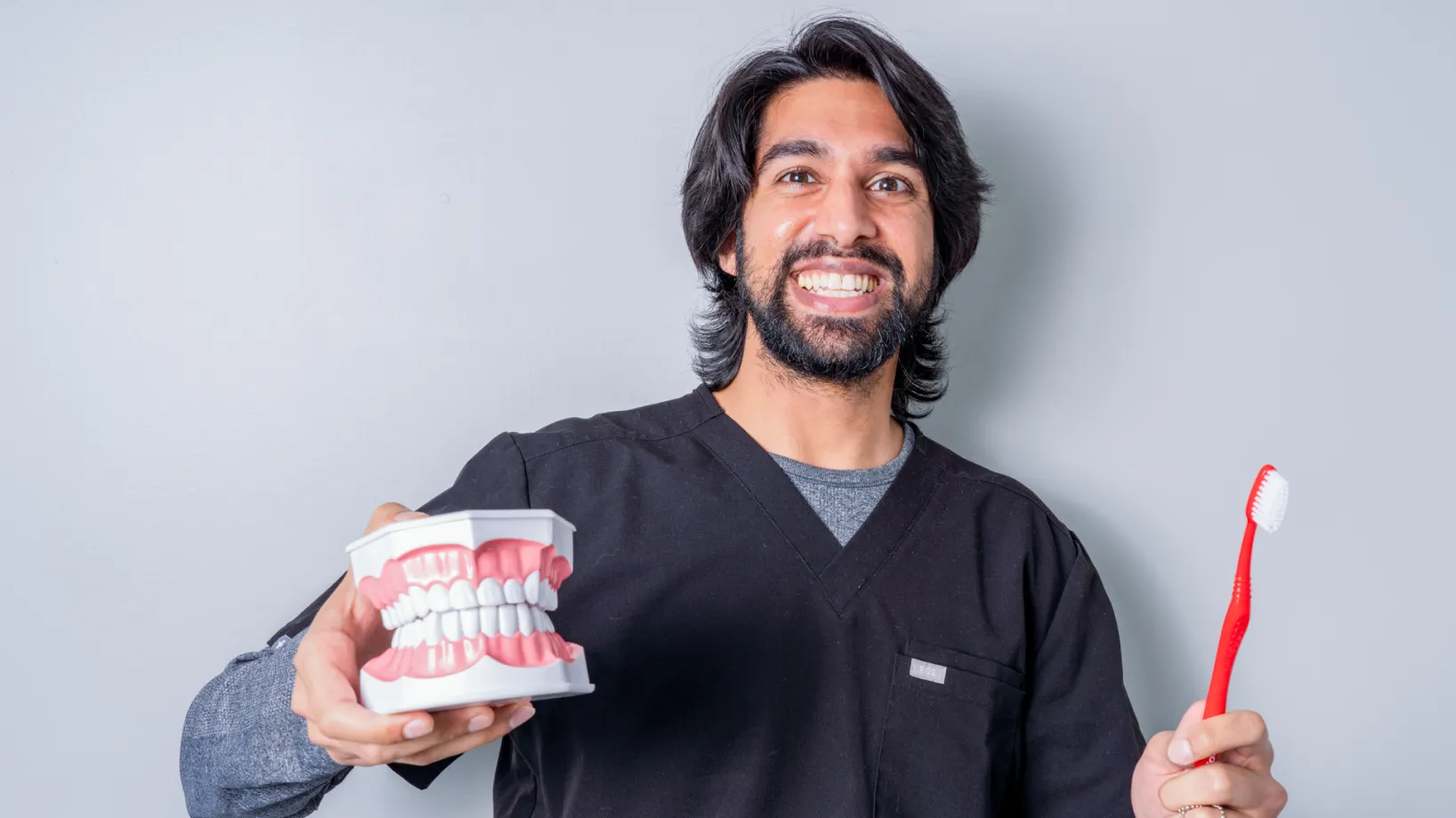 Smiling man in black scrubs holding a dental model and a red toothbrush against a gray background.