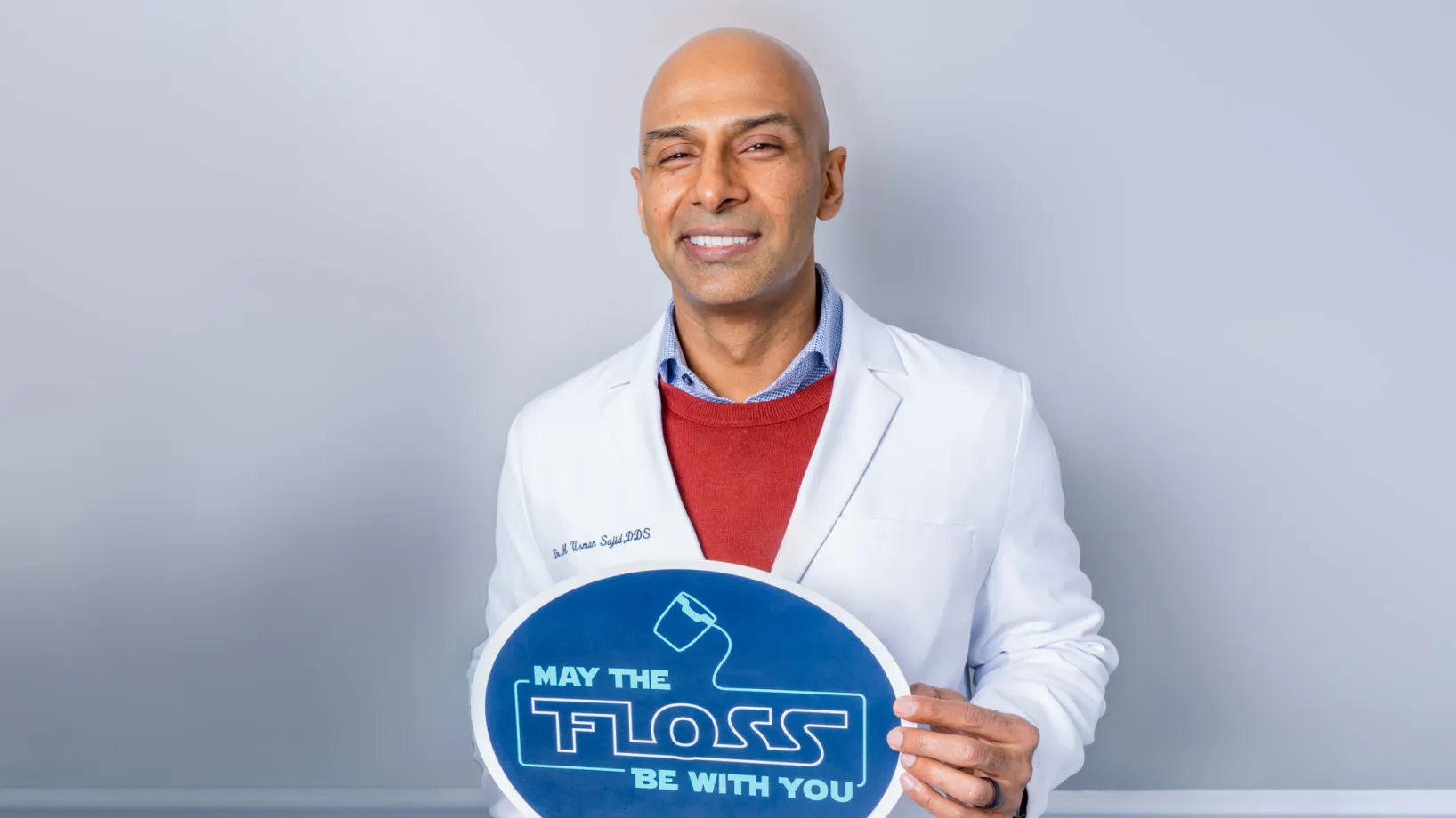Smiling bald male dentist in white coat holding a sign that says 'May the floss be with you'.